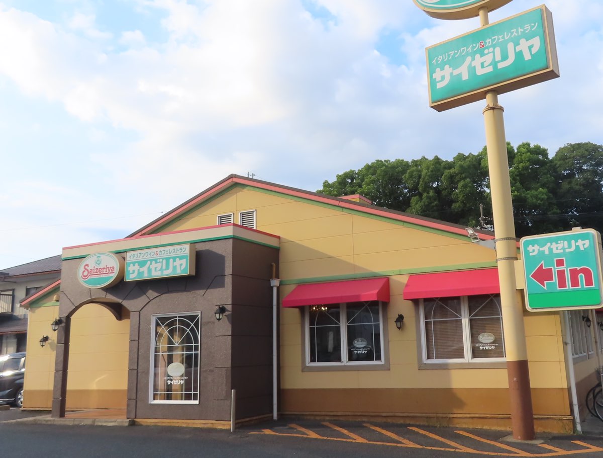 夏の明るい空の下の、平屋のサイゼリヤの店。黄色い外壁に赤い日よけ、入口の上と柱の看板に「サイゼリヤ」の文字、右に「サイゼリヤ in」の矢印の看板がある。人は写っていない。